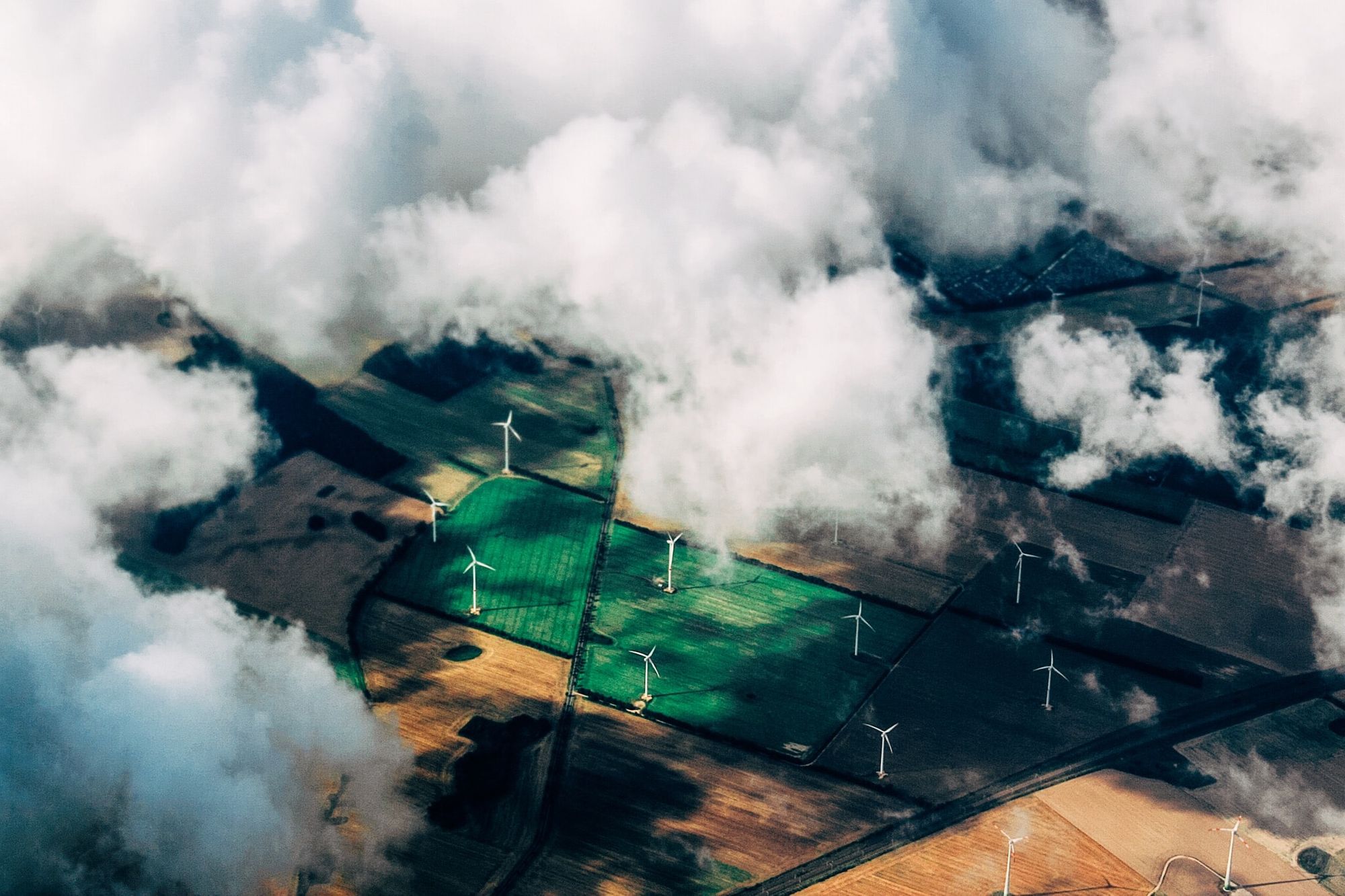 An aerial shot of wind turbines in fields, with fluffy clouds obscuring some of the view