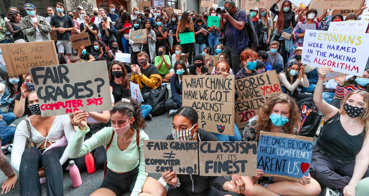 A group of protestors - many of them young people, and many wearing facemasks - standing and sitting. They hold signs that say 'Fair grades? Computer says no', and 'Classism at its finest', and 'No Etonians were harmed in the making of this algorithm'. h.