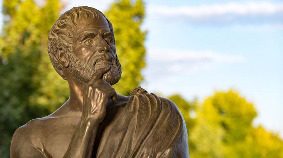 A bronze statue of a bearded person with their finger on their chin in a thinking position, photographed from the chest upwards, with trees and blue skies behind.