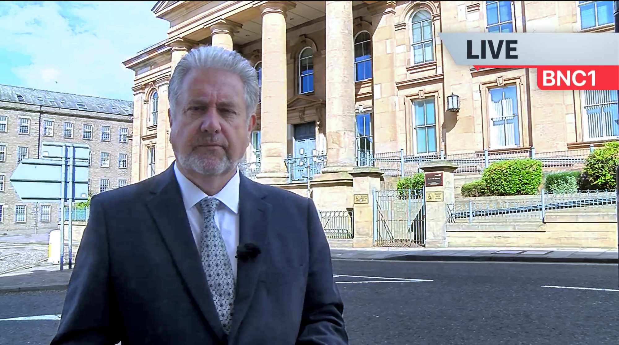Real-life BBC reporter Andrew Anderson presents a news report from digital theatre show The Evidence Chamber outside Dundee Court. Andrew is a white man with neat white hair and beard; he stands in front of an impressive building with four pillars.