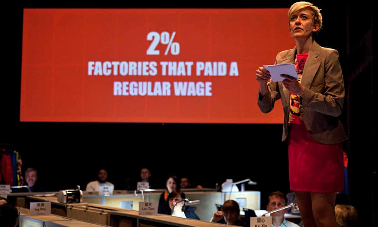 In Metis’ immersive theatre production World Factory, theatre audiences played out the roles of factory owners. In this picture, one of the actors facilitating the piece, a white person wearing a microphone reads notes off a card to the interactive audience. A projection in the background reads ‘2% factories that paid a regular wage’.
