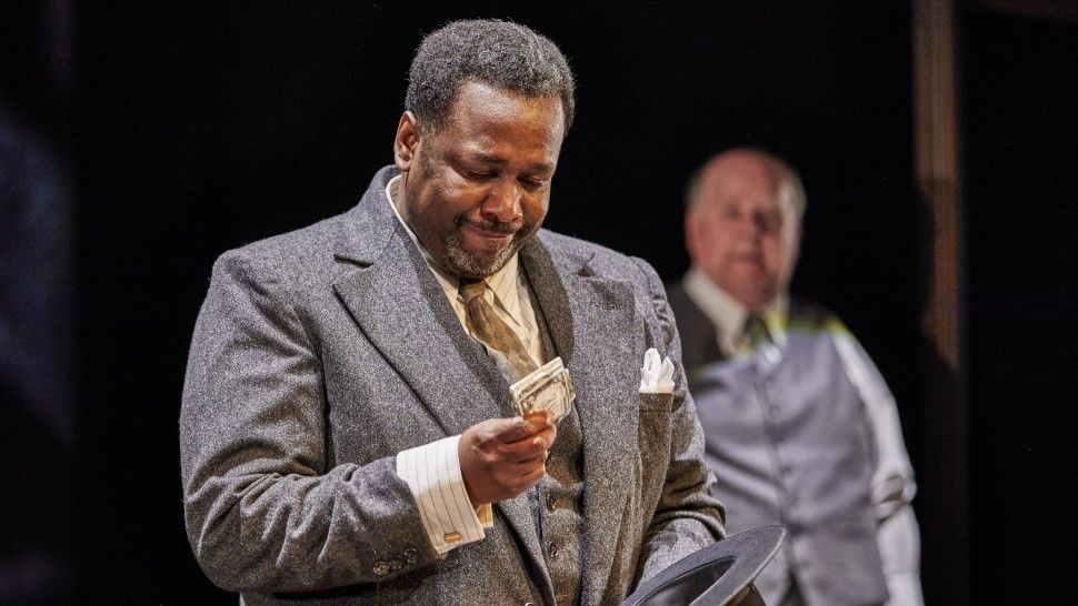 Wendell Pierce playing Willy Loman in Death of a Salesman - an African American man in a grey suit looks at a bundle of banknotes