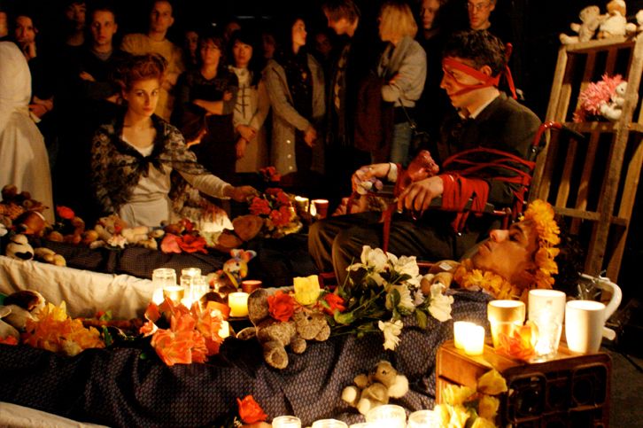 Hotel Medea by ZU-UK, one of the pioneers of the form, and now one of the companies questioning the future of immersive theatre. In the image, a person tied to a wheelchair with red ribbons and a person in a shawl watch over two people lying down. The people lying down are decorated with flowers, lights and soft toys.