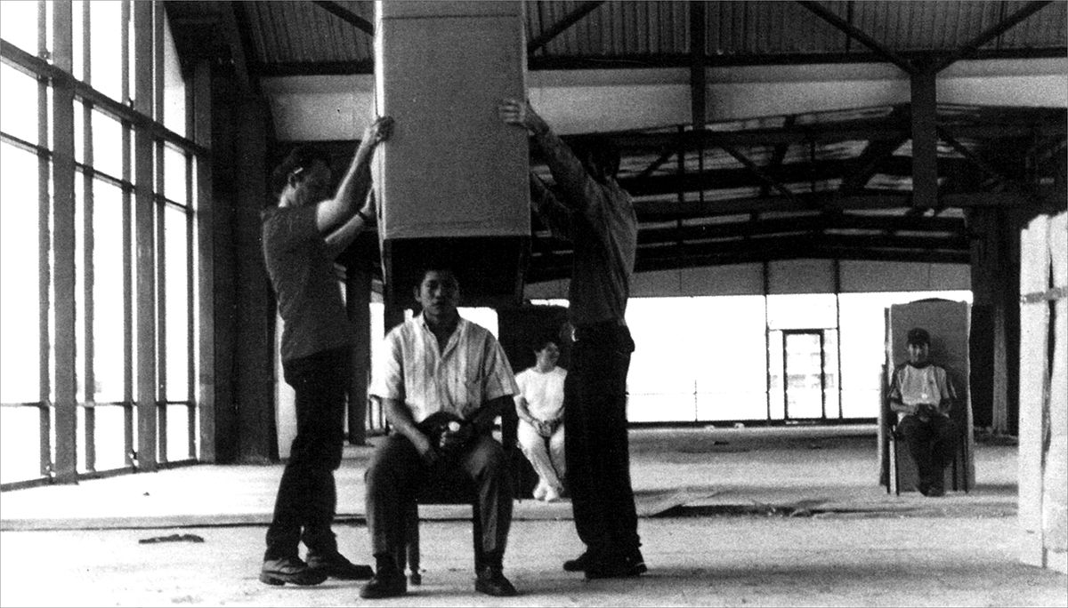 A photo of Workers Who Cannot Be Paid, Remunerated to Remain Inside Cardboard Boxes, showing 3 large boxes and 3 people sitting on chairs, waiting for the box to be lifted over them.