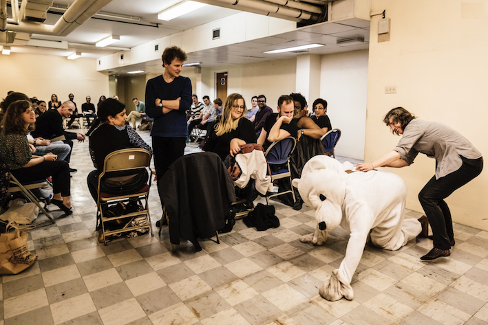 A picture of the performance of The Privileged at Spill Festival. Cuddles, the polar bear, has escaped from the circle of audience members. An audience member is standing next to him, trying to restrain him.