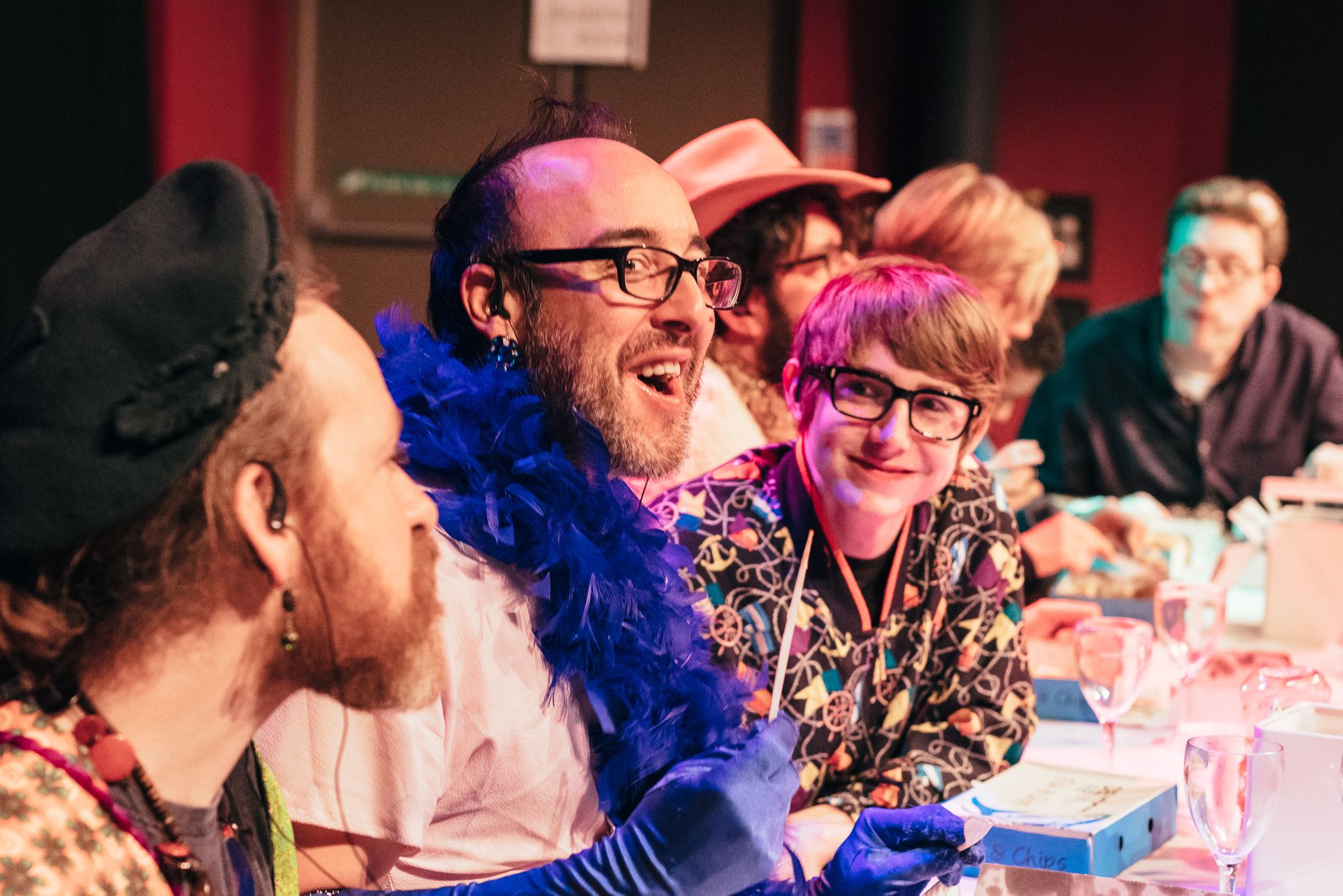 In Fast Familiar’s audience-centred artwork Disaster Party, the interactive audience took on the roles of guests at a party where - you’ve guessed it - everything goes wrong. Here, a white man with stubble wears a blue weather boa and shiny earrings. He is laughing and eating fish and chips. A younger person on his left is looking at him and also laughing.