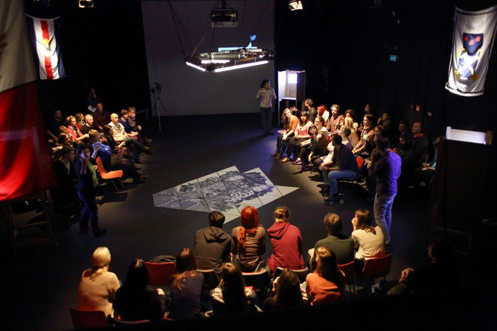 Three groups of 15-20 people are seated on plastic chairs around a map on the floor. Flags hang from the walls above each group. This is a scene from Coney's immersive theatre show, Early Days of a Better Nation.