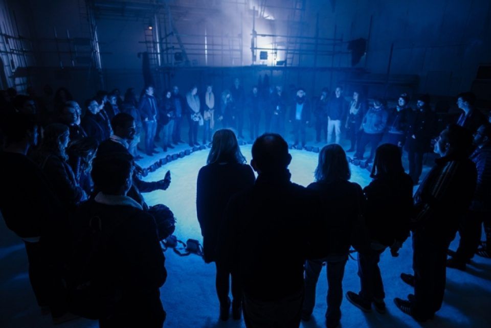 A group of people stand in a circle in a dilapidated building. There is snow on the floor and an eerie blue light. This is a photo from immersive theatre company Raucous's production Ice-Road. pr
