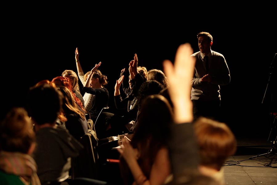 In this scene from immersive theatre production Ten out of Ten, two rows of people sit on chairs. Some of them have their hands raised, as if to answer a question. A white man in a grey v-neck jumper talks to them.