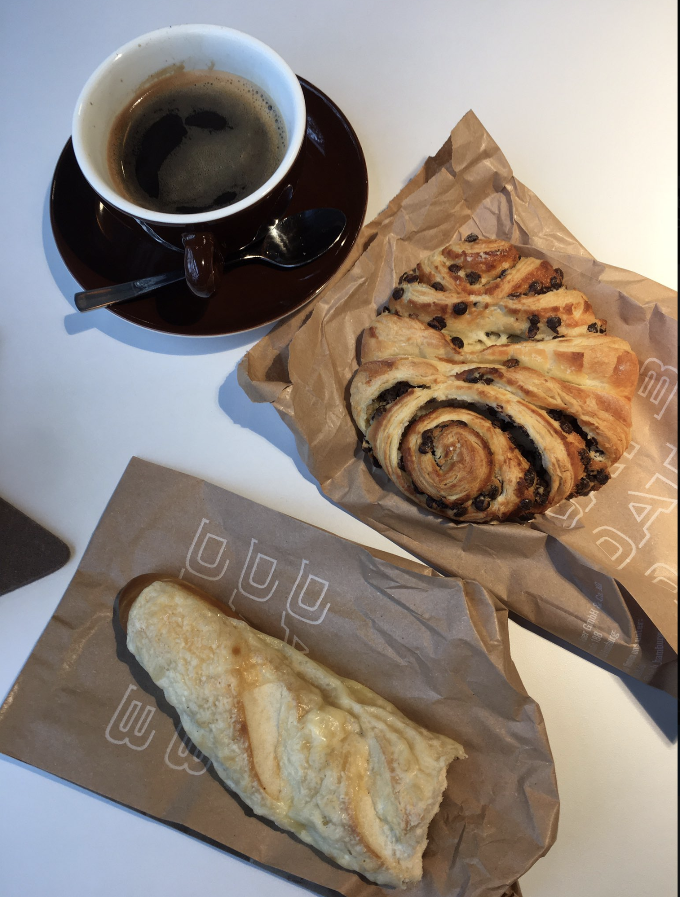 A cup of black coffee sitting on a black saucer and a chocolate filled pastry and half a cheese pastry. The pastries sit on paper bags