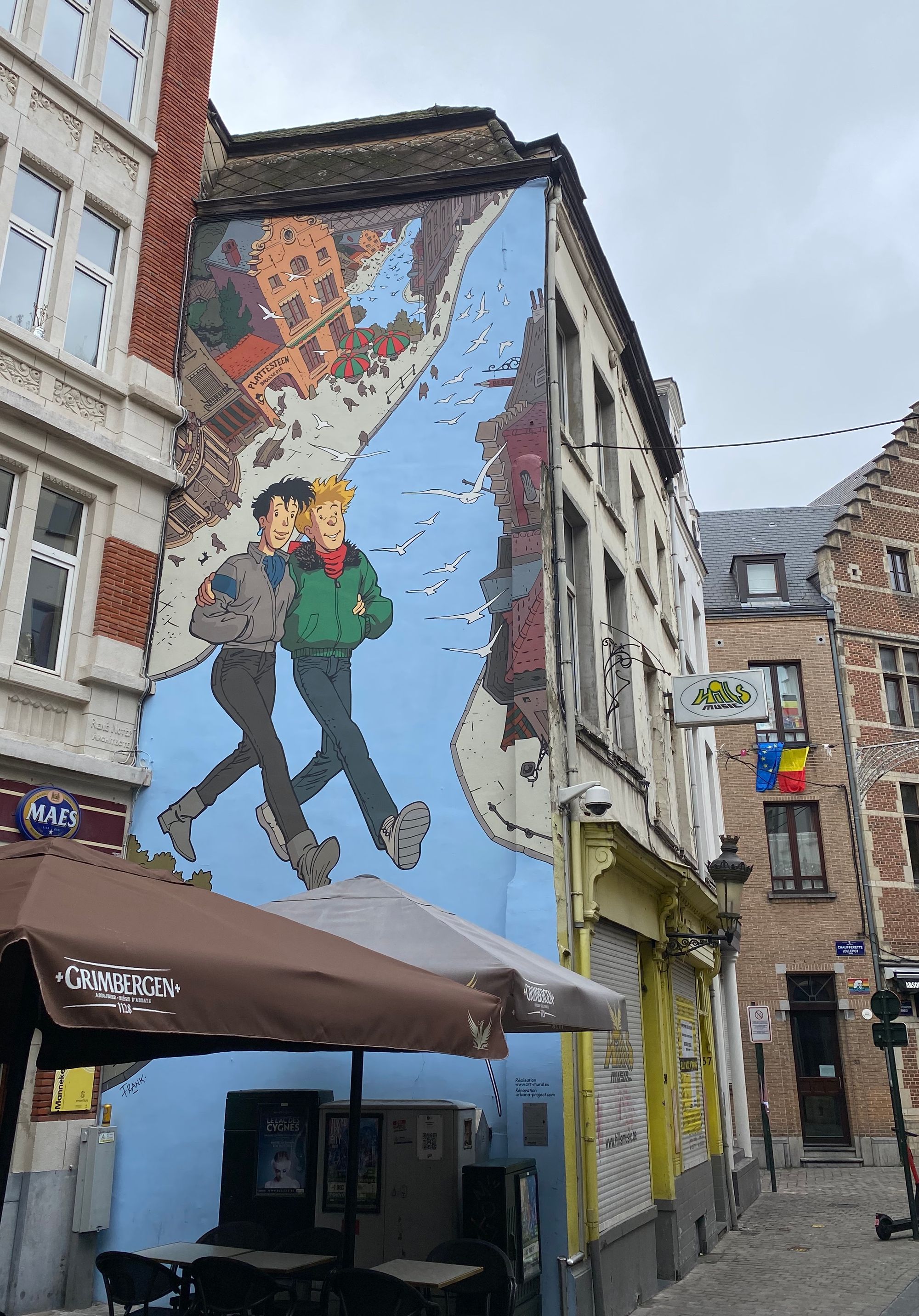 The side of a building which has been painted as cartoon-style street art. The street art shows two people with short hair hugging as they walk along. In front of the street art there is an umbrella outside a bar, with tables and chairs underneath