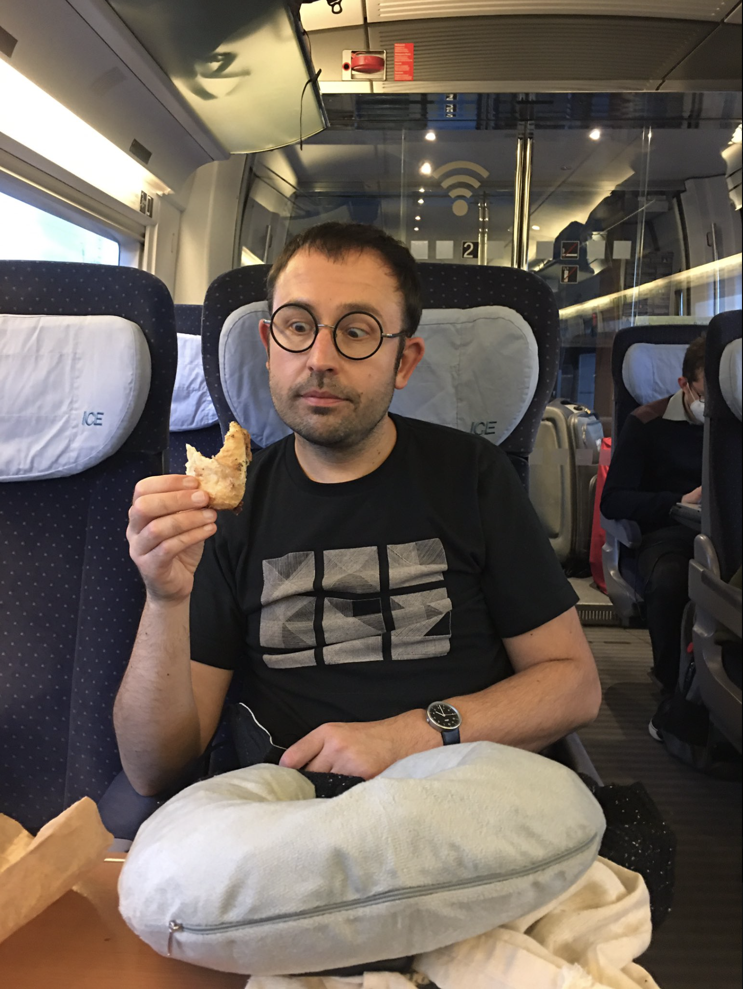 A white man in his 40s, wearing a black t shirt and black glasses is sitting on the seat of a train, looking at a pastry that he is half way through eating. In front of him on the table is a grey neck pillow