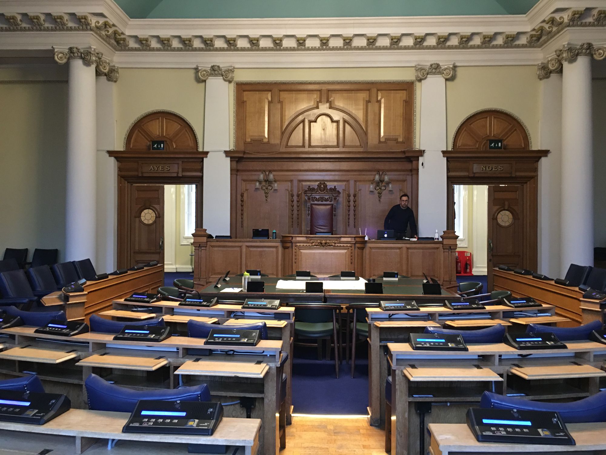 A large, grand-looking civic room. There is lots of wood panelling. At the front of the room are 3 throne-like seats. Framing them are two doors, labelled 'ayes' and 'noes'. In the room are wooden desks and tables.'.