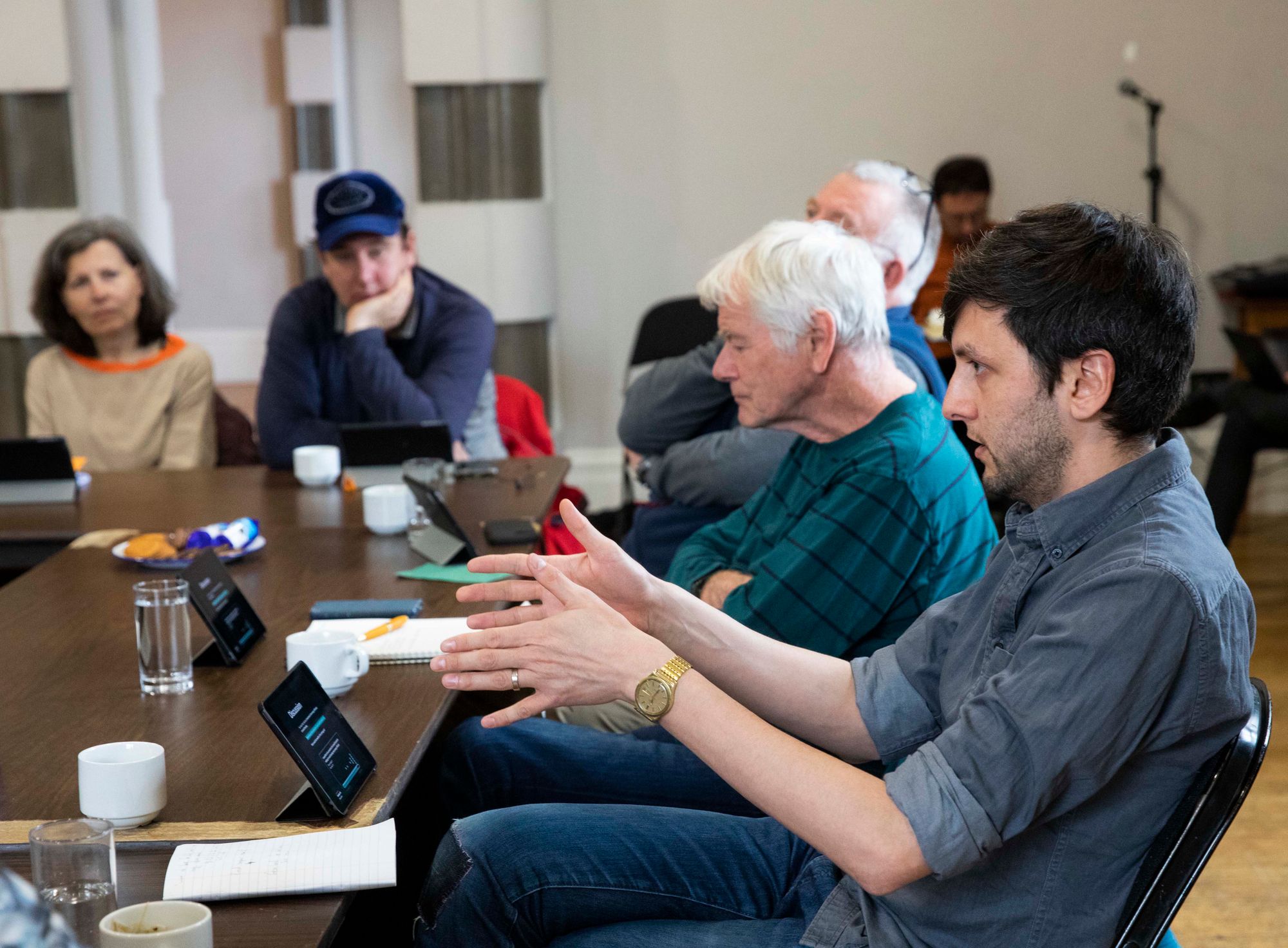 A group of people sit around a table; each person has an ipad. In the foreground, a white person in their 30s is gesturing with their hands to illustrate a point.